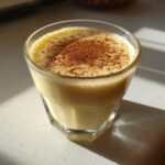 A glass of creamy, pale yellow eggnog topped generously with ground cinnamon, sitting in sunlight.