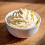 A close-up of thick, creamy homemade mayonnaise swirled in a small white bowl on a wooden surface.