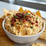 A close-up of creamy million dollar dip served in a white bowl, topped generously with crumbled bacon and chopped green onions.
