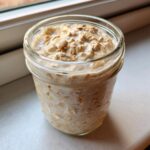 Close-up of creamy overnight oats soaking in milk inside a clear glass mason jar.