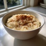 A white bowl filled with creamy rice pudding topped generously with ground cinnamon, set near a sunlit window.