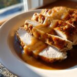 Close-up of sliced, seasoned crockpot turkey breast smothered in rich brown gravy on a white plate.