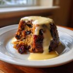 A close-up of a rich, dark slice of plum pudding generously topped with flowing vanilla custard.