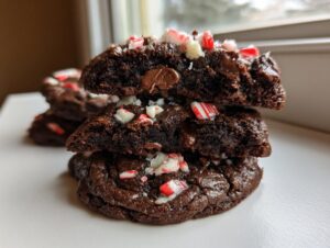 A stack of three rich double chocolate peppermint cookies, with the top two broken open to show the gooey center and peppermint pieces.