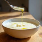 A spoonful of olive oil is drizzled over a bowl of creamy roasted cauliflower soup, garnished with crumbled cauliflower.