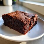A close-up of a rich, fudgy square of date brownies resting on a white plate, highlighted by natural sunlight.