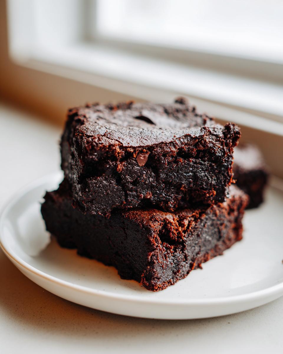Two rich, fudgy date brownies stacked on a small white plate near a bright window.