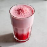 Close-up of a tall glass filled with frothy, layered strawberry milk on a white marble surface.