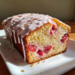 A golden baked loaf of strawberry bread, sliced to show moist interior with visible strawberries, topped with a white glaze.