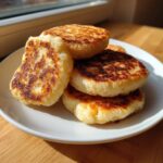 A stack of four golden-brown, pan-fried irish potato farls served on a white plate near a sunny window.