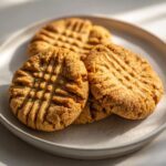 Three golden brown peanut butter cookies with a classic fork crosshatch pattern stacked on a speckled white plate.