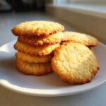 A stack of golden brown, textured butter cookies resting on a white plate, highlighted by sunlight.