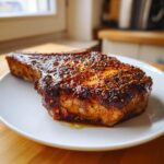 A thick, juicy baked pork chop with a dark, caramelized crust seasoned with visible mustard seeds, resting on a white plate.