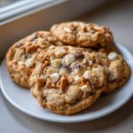 Four freshly baked kitchen sink cookies loaded with white chocolate chips and pretzel pieces piled on a white plate.