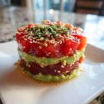A beautiful, stacked presentation of tuna tartare layered with creamy avocado, topped with sesame seeds and herbs.