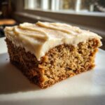 A close-up of a moist slice of applesauce cake topped with thick cream cheese frosting and a dusting of cinnamon.