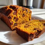 A close-up of moist chocolate chip pumpkin bread, with one slice cut and resting next to the main loaf on a white plate.