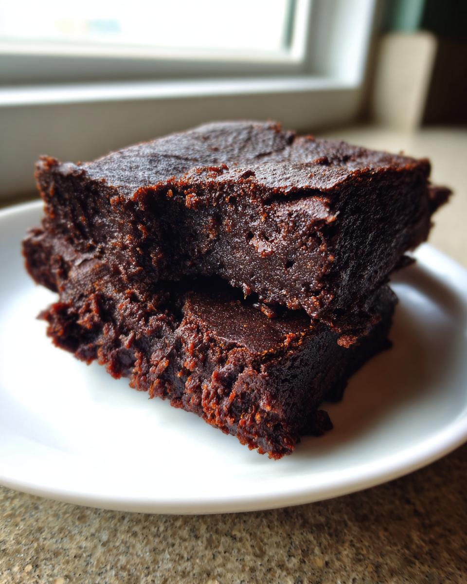 Close-up of two stacked, fudgy date brownies on a white plate near a window.