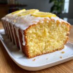 Close-up of a moist, yellow lemon loaf cake with white glaze dripping down the sides and topped with thin lemon slices.