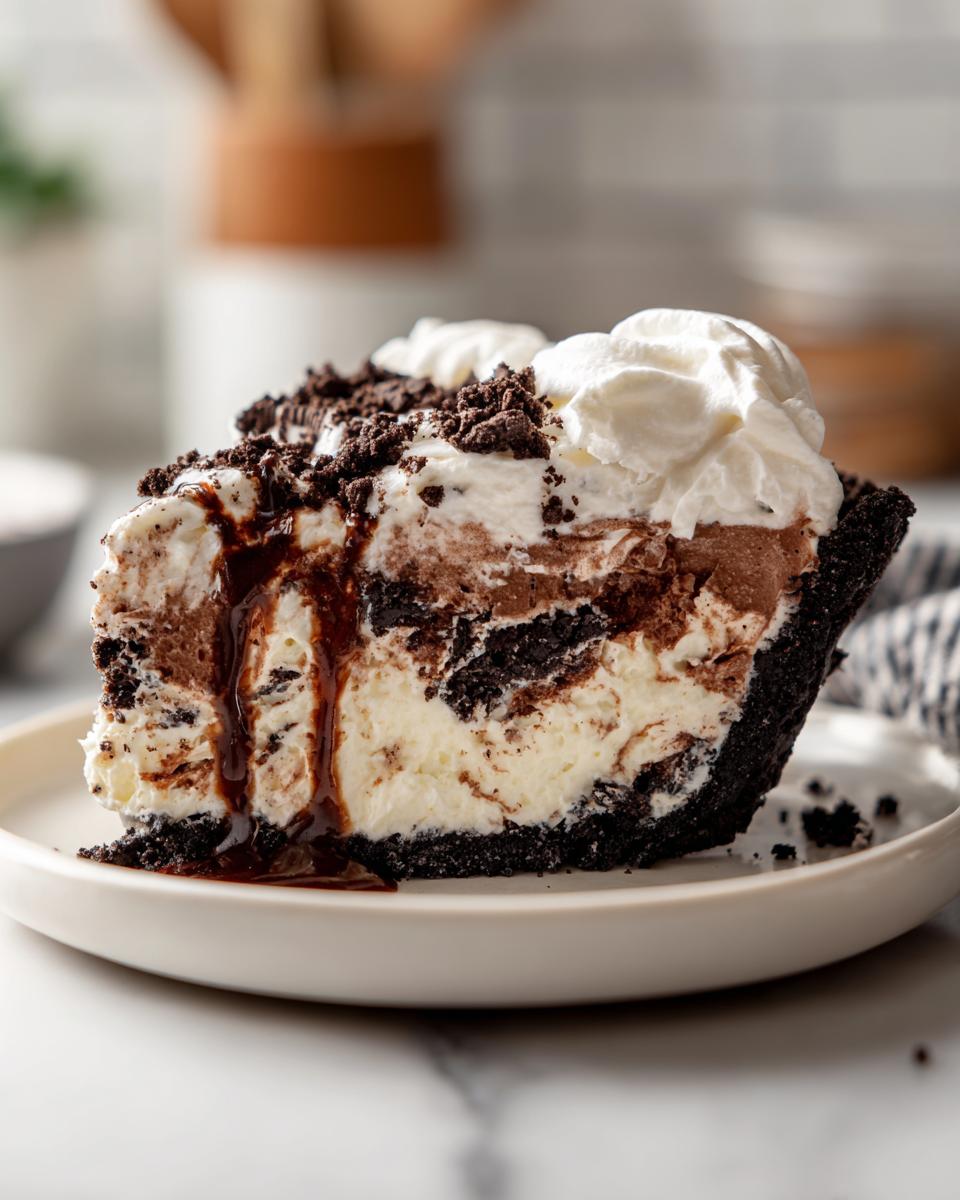 A decadent slice of no-bake ice cream pie featuring layers of vanilla and chocolate filling in a dark crust, topped with whipped cream and chocolate syrup.