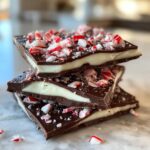 Three stacked squares of homemade peppermint bark recipe showing dark chocolate, white mint layer, and crushed candy canes on top.