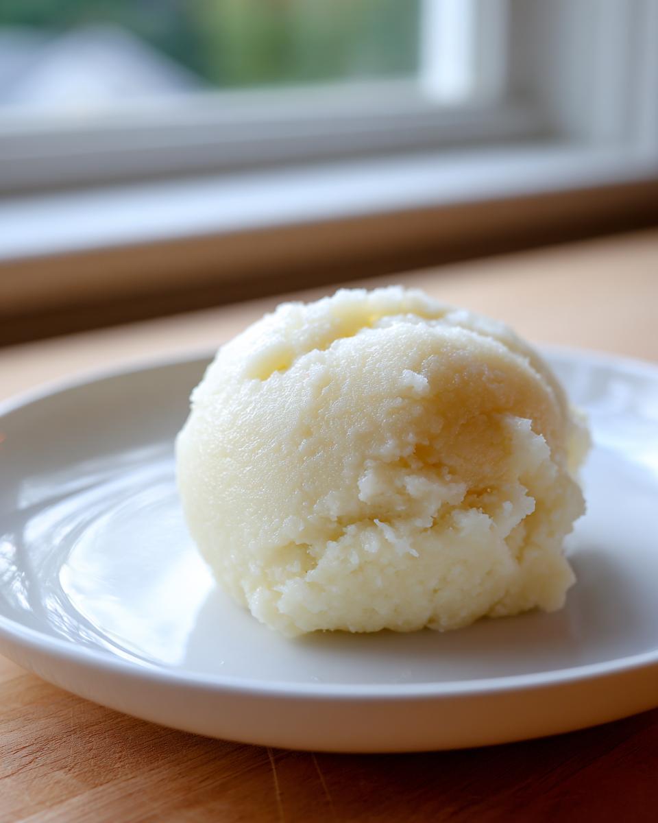 A scoop of smooth, pale yellow fufu served on a white plate, ready to eat.