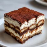 Close-up of a perfect slice of layered tiramisu recipe, dusted heavily with cocoa powder.