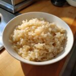 Close-up of perfectly seasoned sushi rice grains glistening in a white bowl on a wooden countertop.