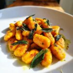 A close-up of vibrant orange sweet potato gnocchi tossed in brown butter sauce with crispy sage leaves.