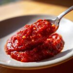 A spoonful of thick, vibrant red pizza sauce, showing small chunks of garlic or herbs, being lifted from a small pile on a light-colored plate.