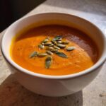 Close-up of a bowl of vibrant orange roasted butternut squash soup topped with pumpkin seeds and sage leaves.