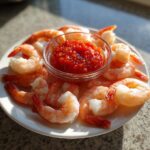 A white plate featuring peeled shrimp cocktail arranged around a small glass bowl of zesty red dipping sauce.