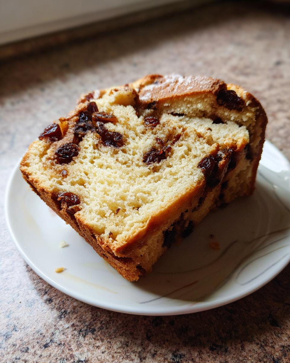 A thick slice of homemade raisin bread showing a soft, yellow crumb packed with dark raisins, served on a small white plate.