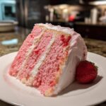 A moist slice of pink strawberry cake with white frosting layers, served with a fresh strawberry.
