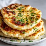 Close-up of a stack of three fluffy naan bread pieces brushed with melted butter and sprinkled with fresh green herbs.