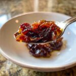 Close-up of a spoonful of rich, dark caramelized onions being lifted from a small white bowl.