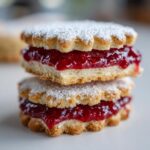 Close-up stack of two delicious linzer cookies filled with bright red raspberry jam and dusted with powdered sugar.
