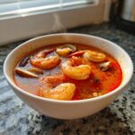 Close-up of a white bowl filled with steaming, vibrant red tom yum soup featuring large shrimp and sliced mushrooms.