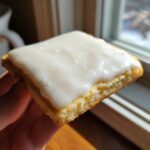 Close-up of a square sugar cookie bar topped with thick, white sugar cookie icing that hardens, held near a window.