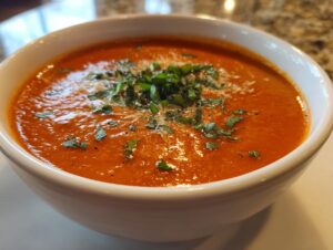 Close-up of a steaming bowl of rich, orange-red tomato basil soup garnished with fresh basil and grated cheese.