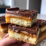 Two stacked cookie bars resembling twix, showing a shortbread base, gooey caramel, and chocolate topping.