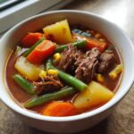 Close-up of a white bowl filled with rich vegetable beef soup, featuring chunks of beef, carrots, potatoes, and green beans.