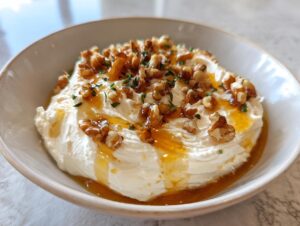 A bowl of creamy, white whipped ricotta dip topped generously with chopped walnuts, honey drizzle, and fresh herbs.