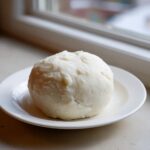 A smooth, white ball of freshly made fufu resting on a small white plate near a window.