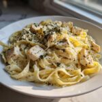 A close-up of a white bowl filled with creamy chicken pasta, topped with cubed chicken and fresh herbs.