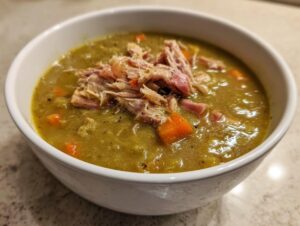 A close-up of a white bowl filled with thick, green pea soup topped with shredded ham and diced carrots.