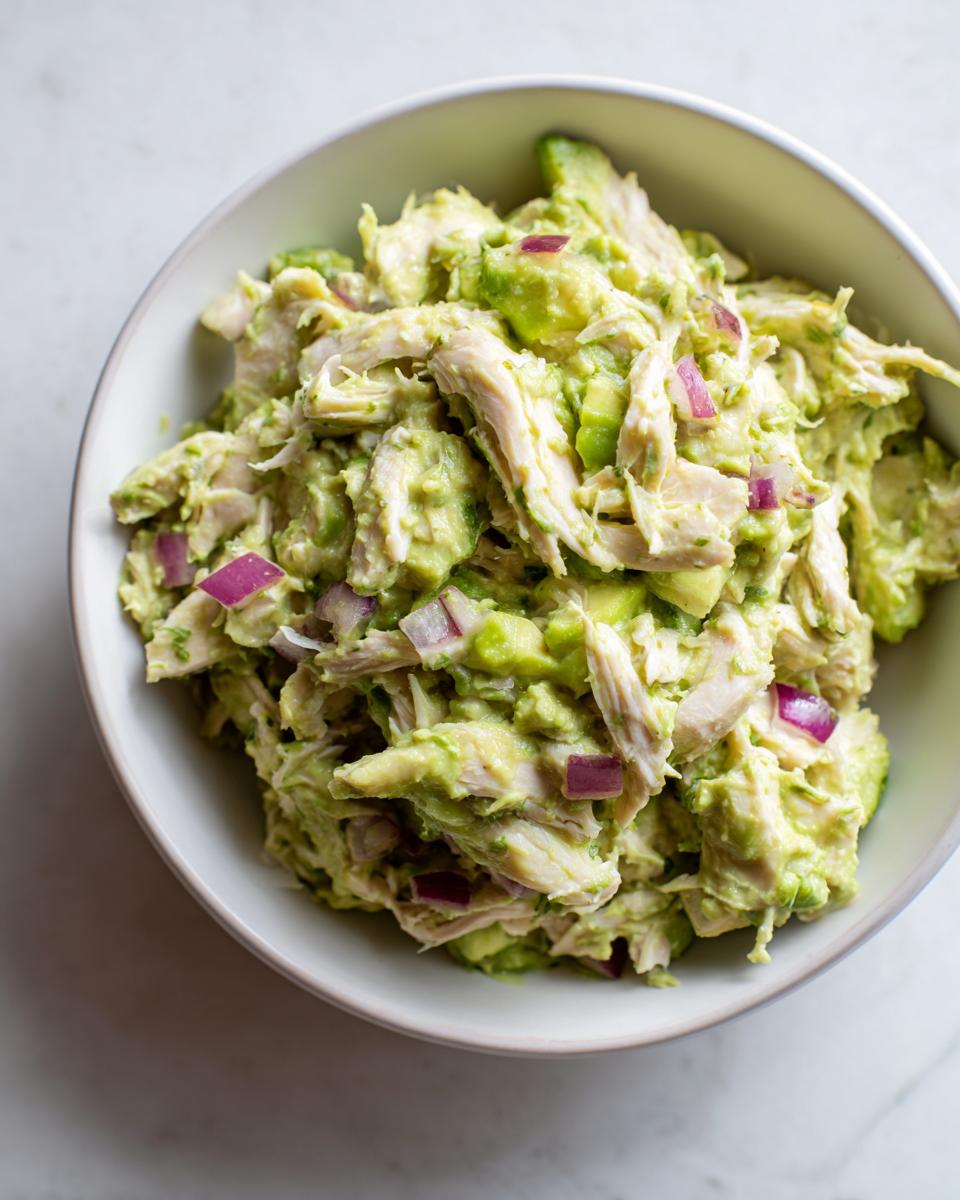 Overhead view of a bowl filled with creamy avocado chicken salad mixed with chunks of avocado and diced red onion.