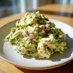 A mound of creamy avocado chicken salad mixed with chunks of chicken and diced red onion, served on a light gray plate.