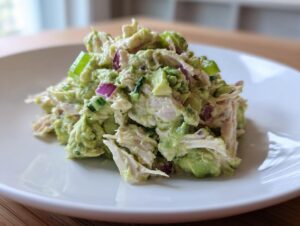 A mound of creamy avocado chicken salad mixed with chunks of red onion and green celery served on a white plate.