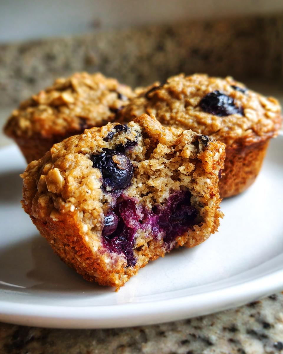 A close-up of a broken blueberry oatmeal muffin showing moist interior and burst blueberries, with two more muffins in the background.